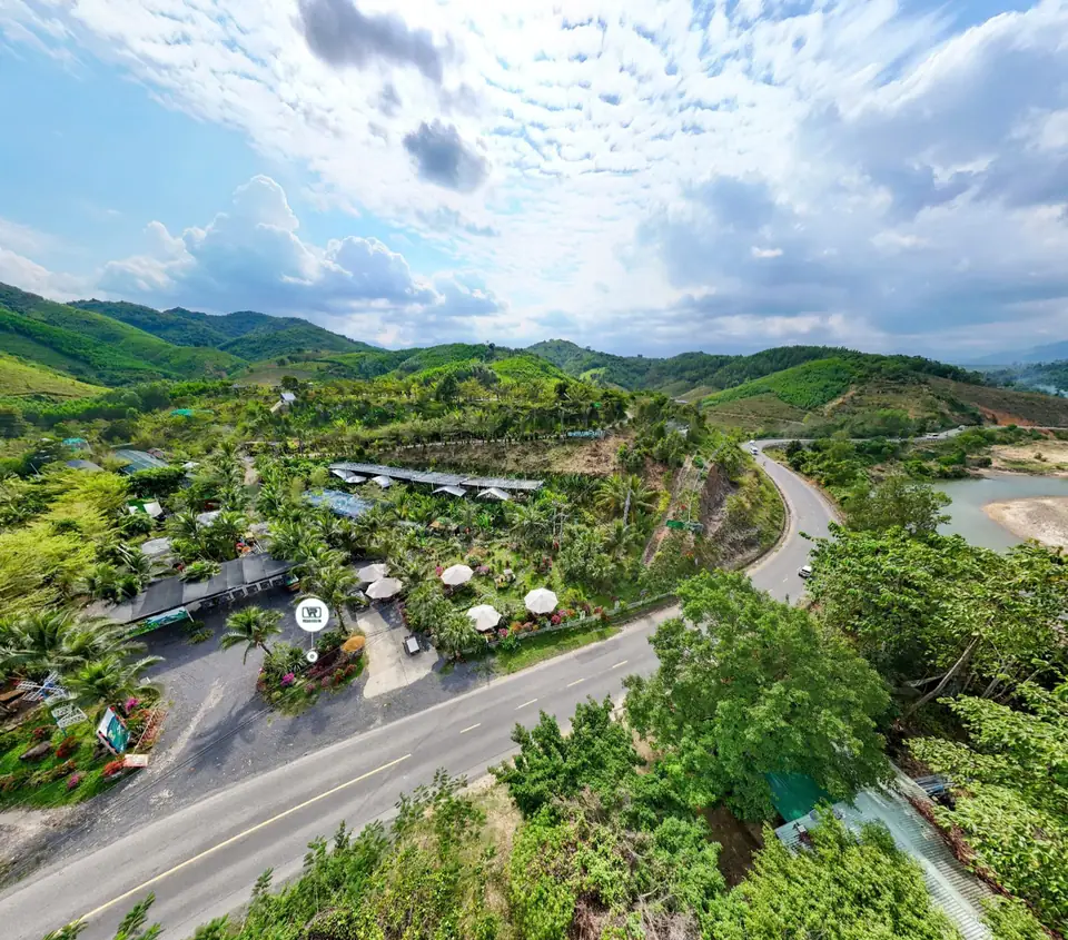 Flycam: quốc lộ 27C chạy trước làng, sông và bãi cát bên phải, đồi xanh và mây phía sau